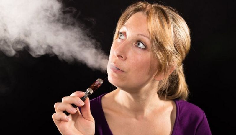 Frau, lila Shirt, blonde, zurückgebundene Haare, schwarzer Hintergrund, raucht E-Zigarette und schaut dabei schräg nach oben