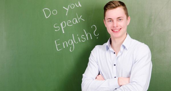 Gymnasiast, ca. 17, 18 Jahre, helles Hemd, lachend, verschränkte Arme, mit dem Rücken zu eine Schultafel, auf der zu lesen ist "Do you speak English?"