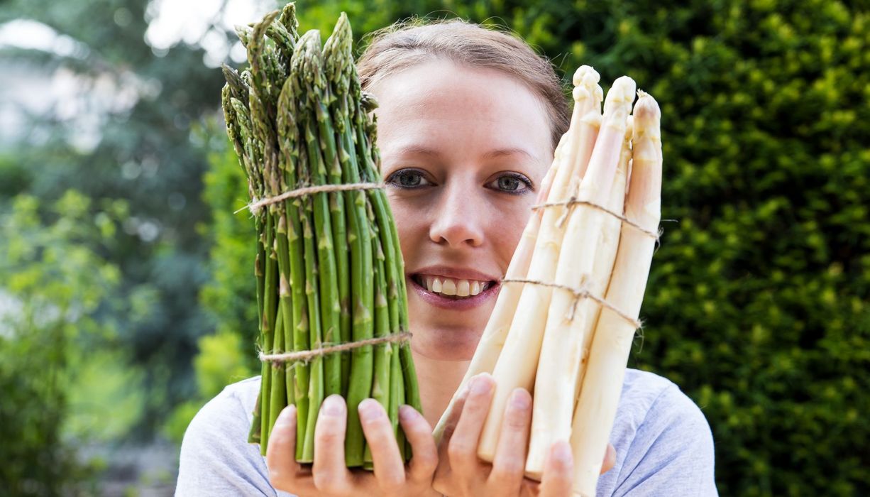 Junge Frau hält je ein Bündel grünen und weißen Spargel in die Höhe. 