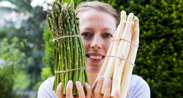 Junge Frau hält je ein Bündel grünen und weißen Spargel in die Höhe. 