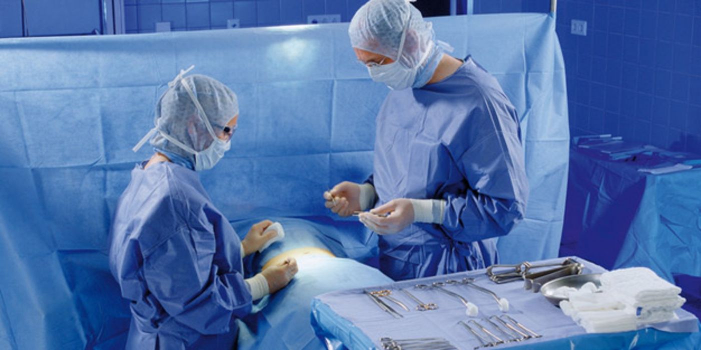 Blick in einen OP-Saal: Zwei Ärzte operieren Patienten, der mit blauen Tüchern abgedeckt ist, im Vordergrund ein Tisch mit OP-Besteck
