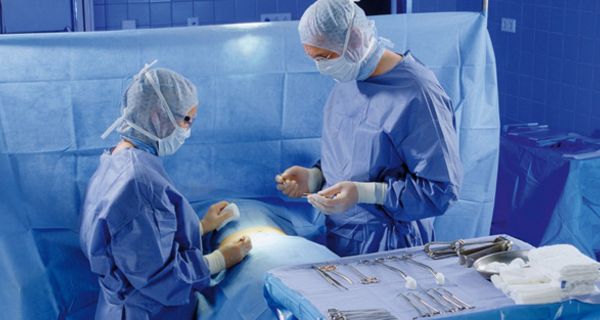Blick in einen OP-Saal: Zwei Ärzte operieren Patienten, der mit blauen Tüchern abgedeckt ist, im Vordergrund ein Tisch mit OP-Besteck