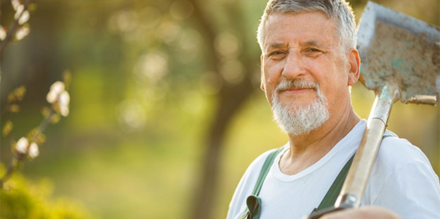 Natur: Mann in den 60ern, grauer Bart und Haare, grüne Arbeitslatzhose, weißes T-Shirt, Spaten über linker Schulter