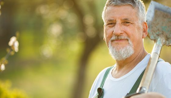 Natur: Mann in den 60ern, grauer Bart und Haare, grüne Arbeitslatzhose, weißes T-Shirt, Spaten über linker Schulter