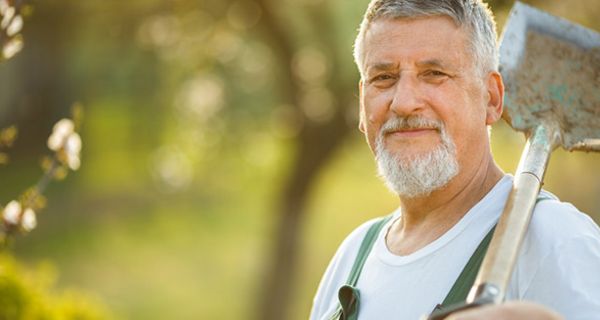 Natur: Mann in den 60ern, grauer Bart und Haare, grüne Arbeitslatzhose, weißes T-Shirt, Spaten über linker Schulter