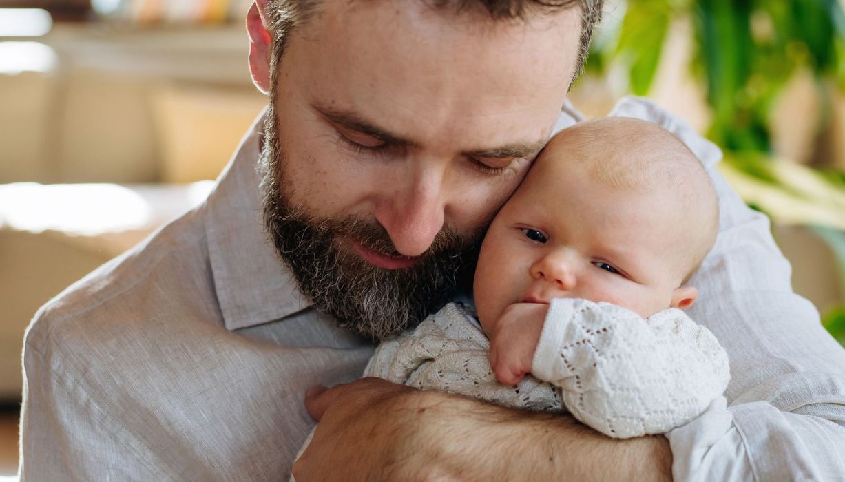 Mann mit einzelnen, grauen Strähnen im Bart, hält ein Baby im Arm.