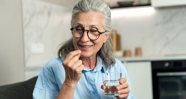 Eine ältere Frau mit einem Glas Wasser in der Hand nimmt eine Tablette und lächelt.