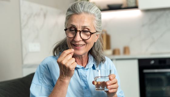 Eine ältere Frau mit einem Glas Wasser in der Hand nimmt eine Tablette und lächelt.