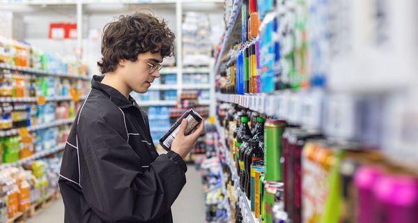 Jugendlicher, steht im Supermarkt vor einem Regal mit Getränken und hält eine Dose Energy Drink in der Hand.