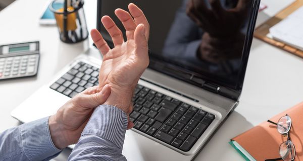 Mann hält sein Handgelenk, im Hintergrund ein Laptop.