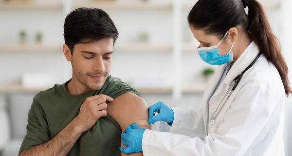 Eine Ärztin klebt einem Mann, wohl nach einer Impfung, ein Pflaster auf den Oberarm.