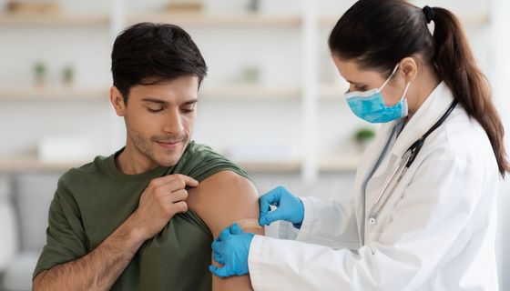 Eine Ärztin klebt einem Mann, wohl nach einer Impfung, ein Pflaster auf den Oberarm.