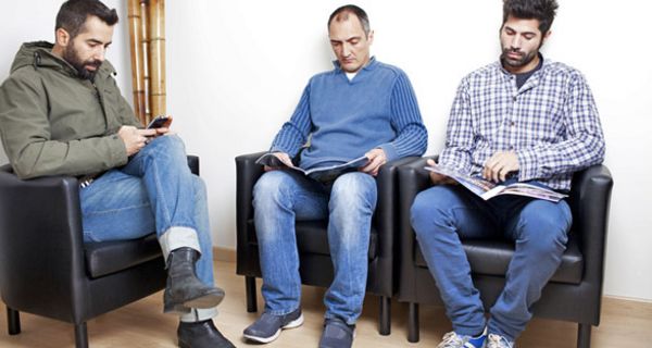 Drei junge Männer in einem Wartezimmer; einer spielt an seinem Handy, die anderen zwei lesen