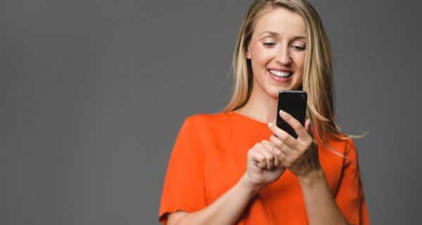 Jüngere Frau mit langen blonden Haaren und weitem orangem Shirt schaut lachend auf ein Smartphone und tippt mit einem Finger darauf. Hintergrund mittelgrau