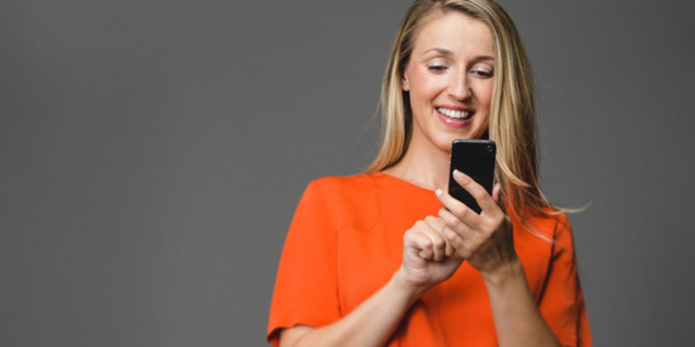 Jüngere Frau mit langen blonden Haaren und weitem orangem Shirt schaut lachend auf ein Smartphone und tippt mit einem Finger darauf. Hintergrund mittelgrau