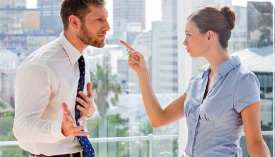 Frau und Mann im Büro vor Fenster stehend im Streitgespräch. Sie zeigt mit dem Zeigefinger auf ihn, er verteidigt sich