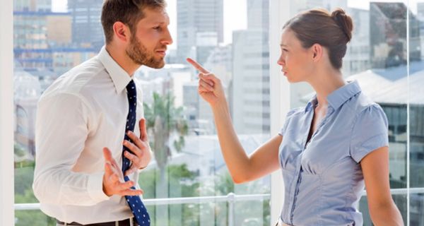 Frau und Mann im Büro vor Fenster stehend im Streitgespräch. Sie zeigt mit dem Zeigefinger auf ihn, er verteidigt sich