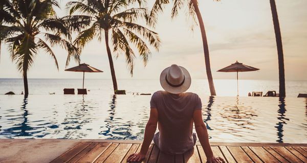 Mann mit Hut, sitzt vor einem Pool mit Blick auf das Meer.