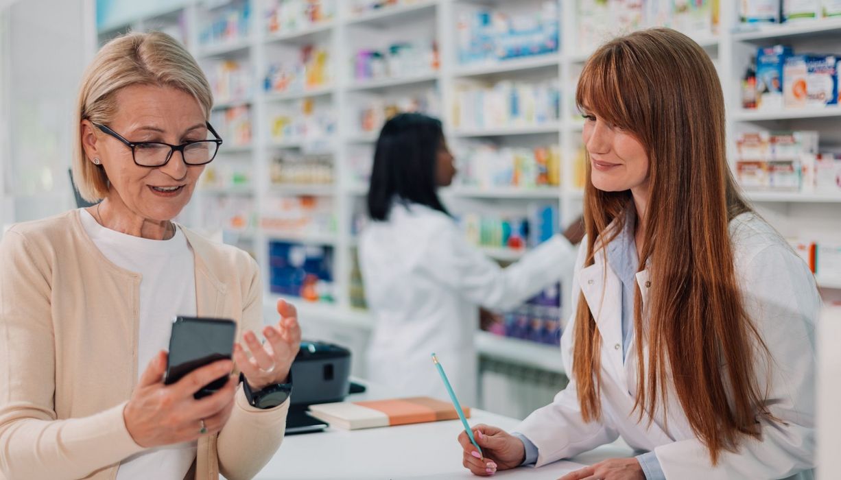 Apothekerin macht sich Notizen, während ihre Kundin ihr etwas mithilfe ihres Smartphones erklärt.