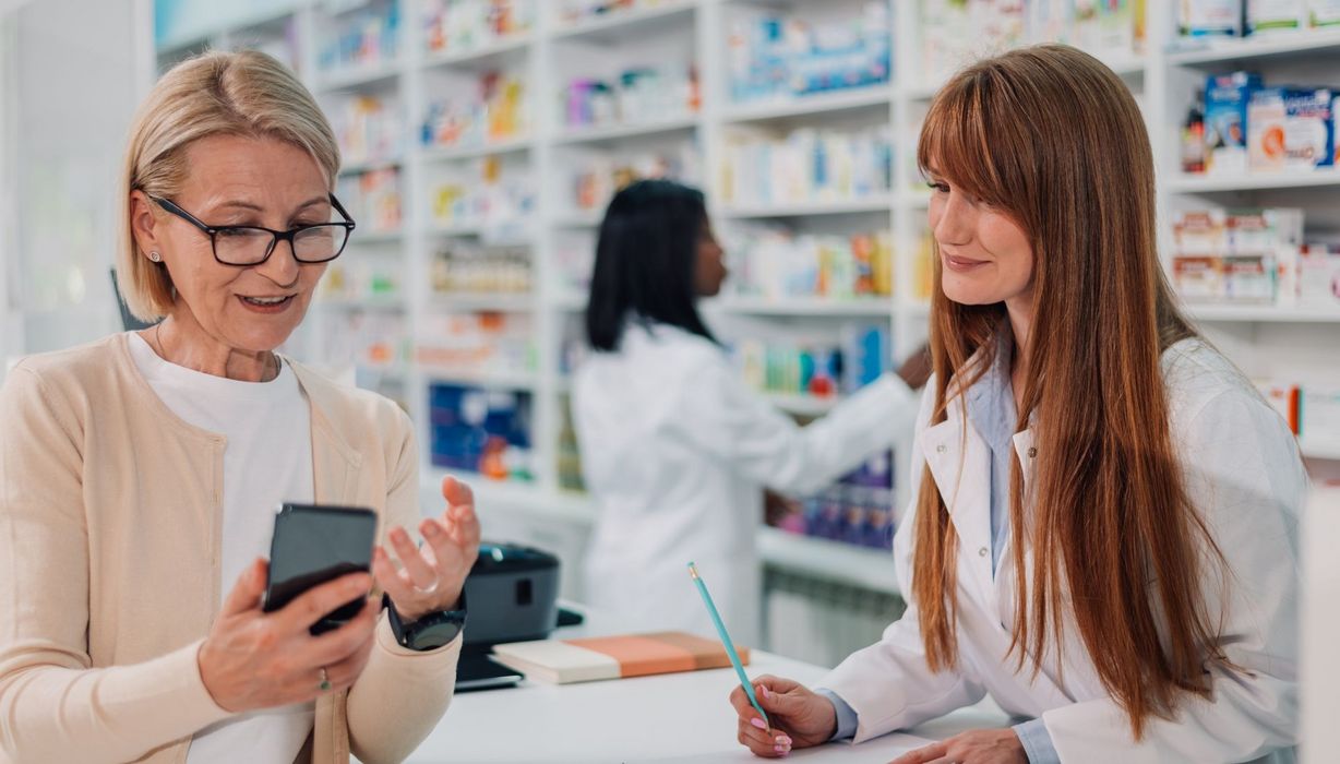 Apothekerin macht sich Notizen, während ihre Kundin ihr etwas mithilfe ihres Smartphones erklärt.