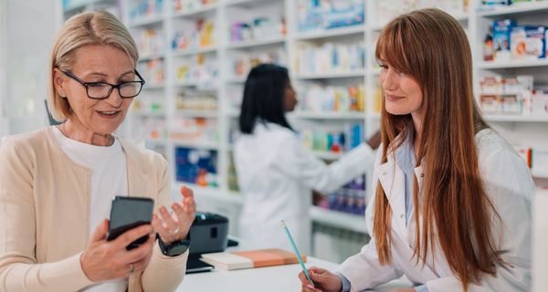 Apothekerin macht sich Notizen, während ihre Kundin ihr etwas mithilfe ihres Smartphones erklärt.
