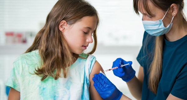 Ein junges Mädchen im grünen T-Shirt wird von einer Frau mit Maske und Handschuhen geimpft.