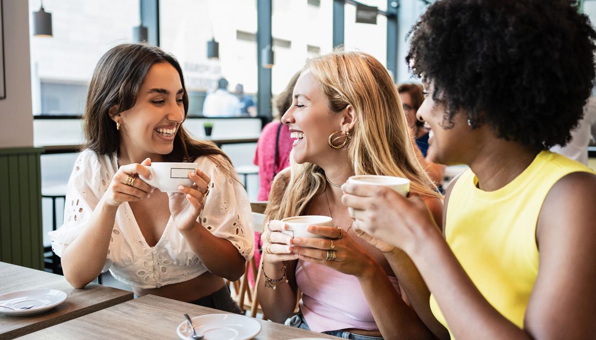 3 junge Frauen sitzen lächelnd mit Kaffee in der Hand in einem Cafe