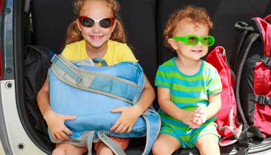 Mädchen und Junge mit Sonnenbrille sitzen im offenen Kofferraum eines Autos nebst Koffern