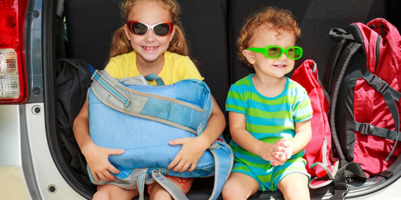 Mädchen und Junge mit Sonnenbrille sitzen im offenen Kofferraum eines Autos nebst Koffern