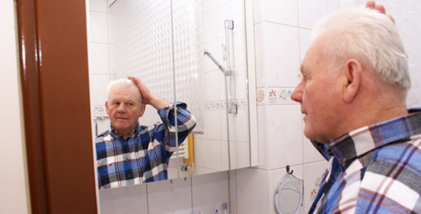 Senior im Holzfällerhemd steht im Bad vor dem Spiegel und fasst sich mit einer Hand an den Kopf