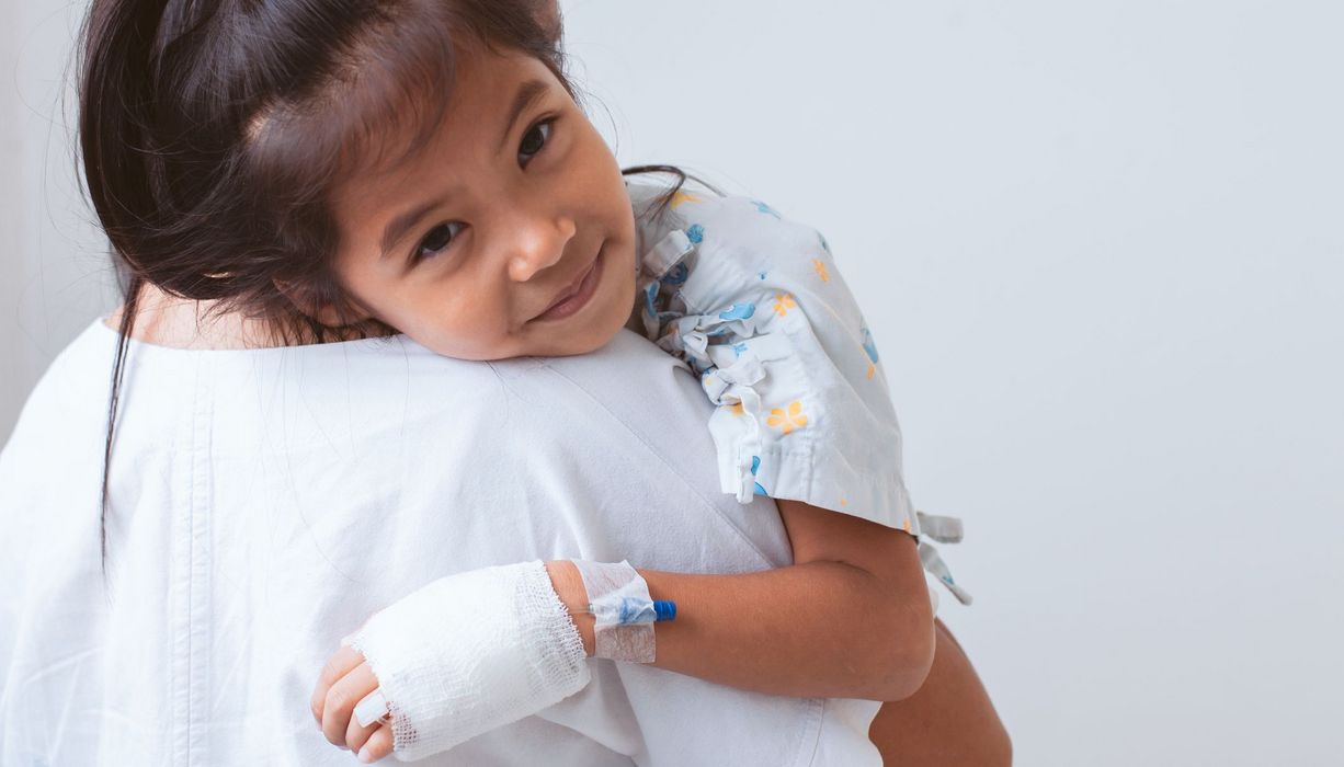 Kind mit Verband um die Hand, liegt im Arm eines Erwachsenen und lächelt.
