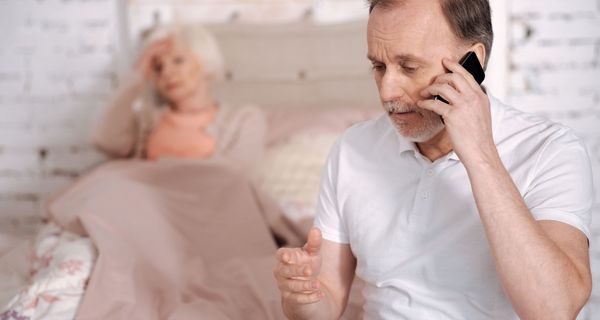 Mann telefoniert, im Hintergrund liegt eine Frau im Bett.