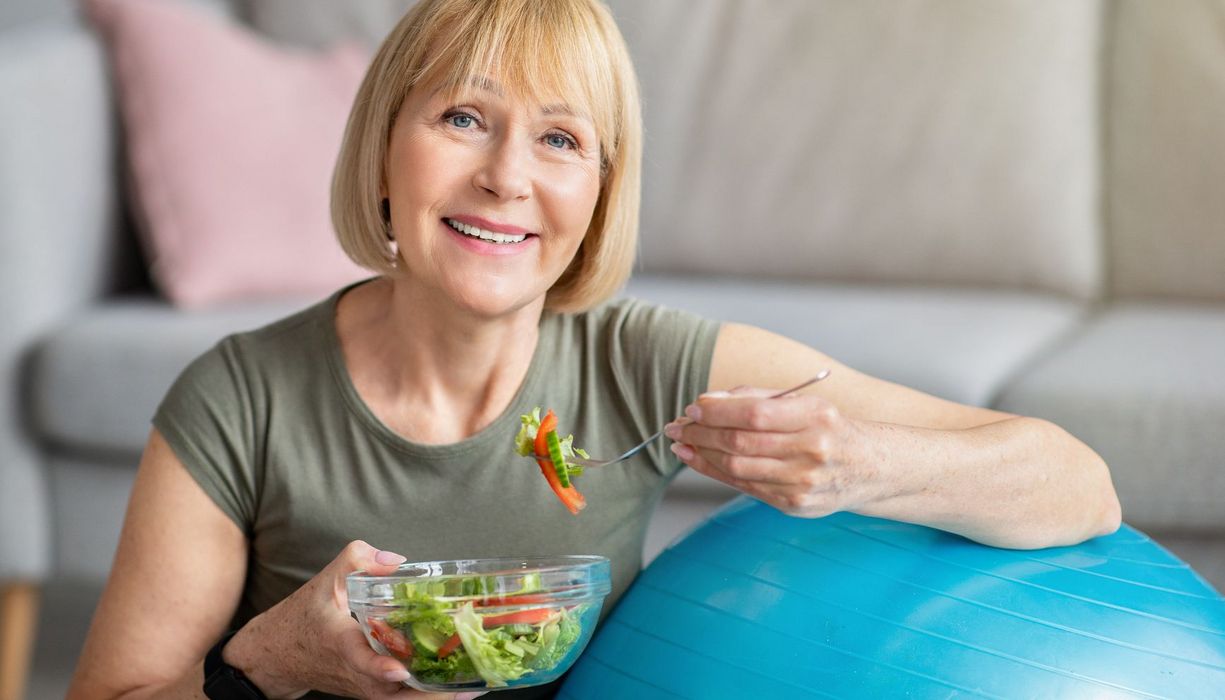 Frau zwischen 40 und 60 isst einen Salat aus einer Glasschüssel, während sie sich an einen Gymnastikball lehnt.