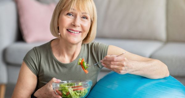 Frau zwischen 40 und 60 isst einen Salat aus einer Glasschüssel, während sie sich an einen Gymnastikball lehnt.