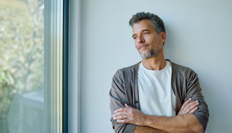 Mann mit grauen Haaransätzen lehnt zufrieden an der Wand und schaut aus dem Fenster.