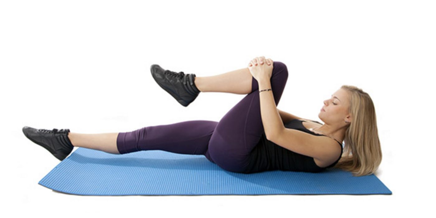 Jüngere Frau auf Gymnastikmatte auf dem Rücken, linkes Knie mit Händen umfassend heranziehend, anderes Bein ausgestreckt, Kopf leicht angehoben