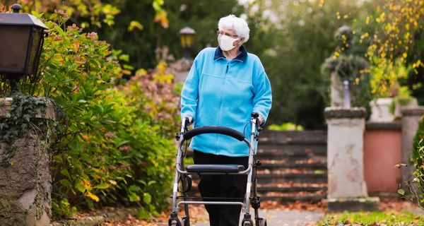 Ältere Frau mit Maske, geht draußen am Rollator spazieren.