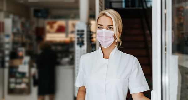 Junge Apothekerin mit Mundschutz steht vor der Apotheke.