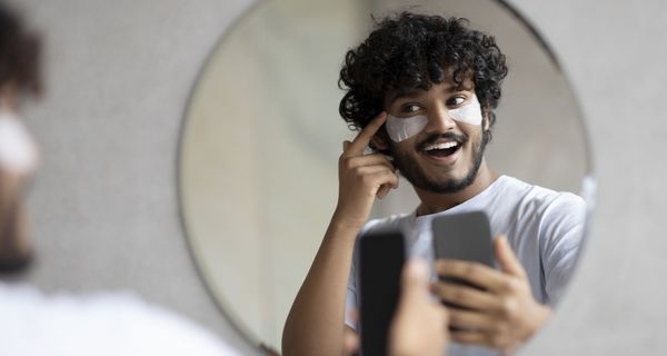 Junger Mann hat sein Handy in der Hand und blickt in den Spiegel. Auf seinen Wangen ist deutlich weiße Kosmetik zu sehen.