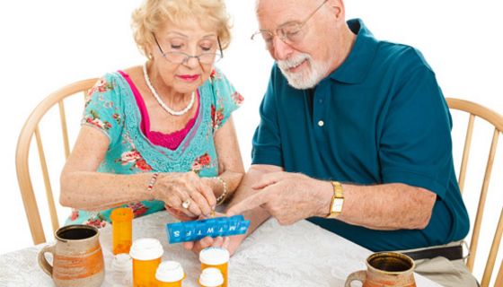 Farbenfroh gekleidetes Seniorenpaar am Tisch bei der Einteilung der Wochenration Tabletten in eine Dosierhilfe
