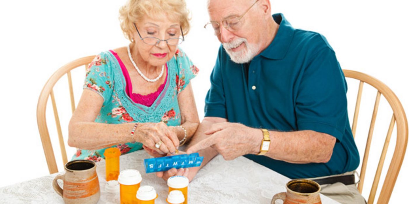 Farbenfroh gekleidetes Seniorenpaar am Tisch bei der Einteilung der Wochenration Tabletten in eine Dosierhilfe