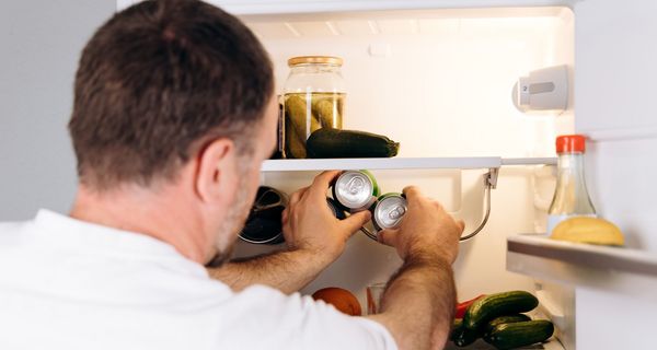 Ein Mann mittleren Alters steht vor einem geöffnetem Kühlschrank und greift zwei Dosen.