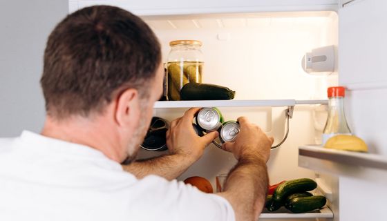 Ein Mann mittleren Alters steht vor einem geöffnetem Kühlschrank und greift zwei Dosen.