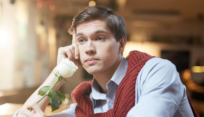 Junger Mann wartet zweifelnd auf sein Date – wohl schon länger. In einer Hand hält er eine Rose.