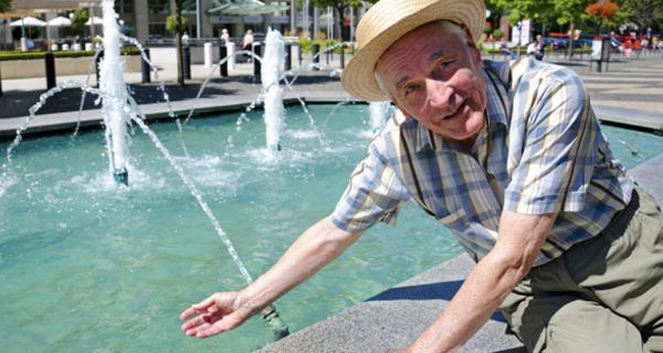 Sommerszene: Senior mit Strohhut an Stadtbrunnen in Bratislava, am Rand sitzend, eine Hand im Wasser