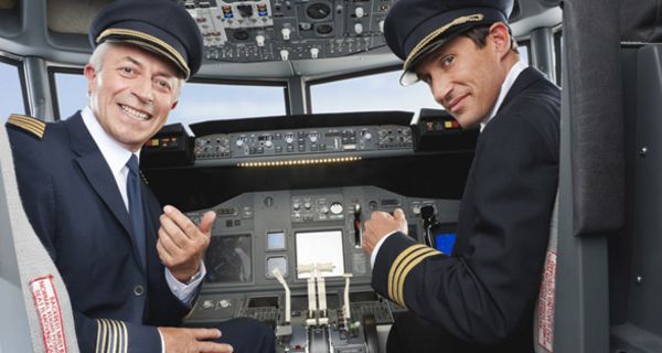 Zwei Piloten im Cockpit schauen aus ihren Sitzen heraus über die Schulter in die Kamera und lächeln