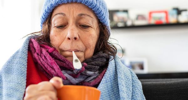 Ältere Frau mit Tee, Mütze und Fieberthermometer im Mund.