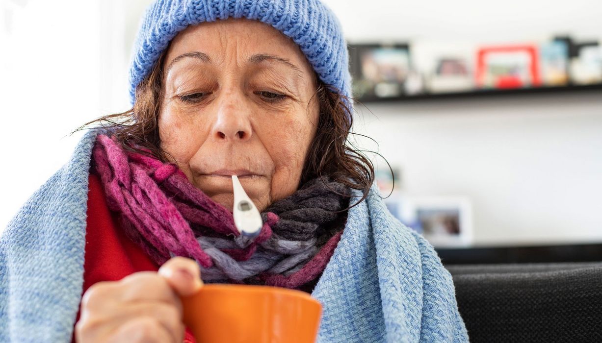 Ältere Frau mit Tee, Mütze und Fieberthermometer im Mund.