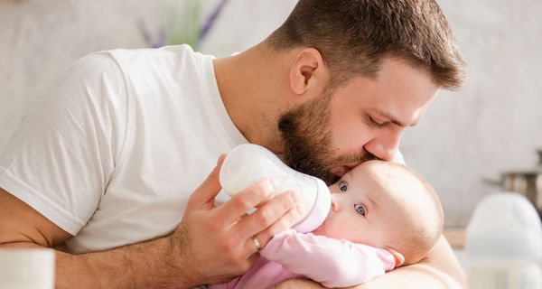 Mann küsst ein Baby auf den Kopf und füttert es mit einem Fläschchen.