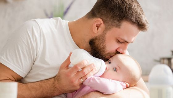 Mann küsst ein Baby auf den Kopf und füttert es mit einem Fläschchen.